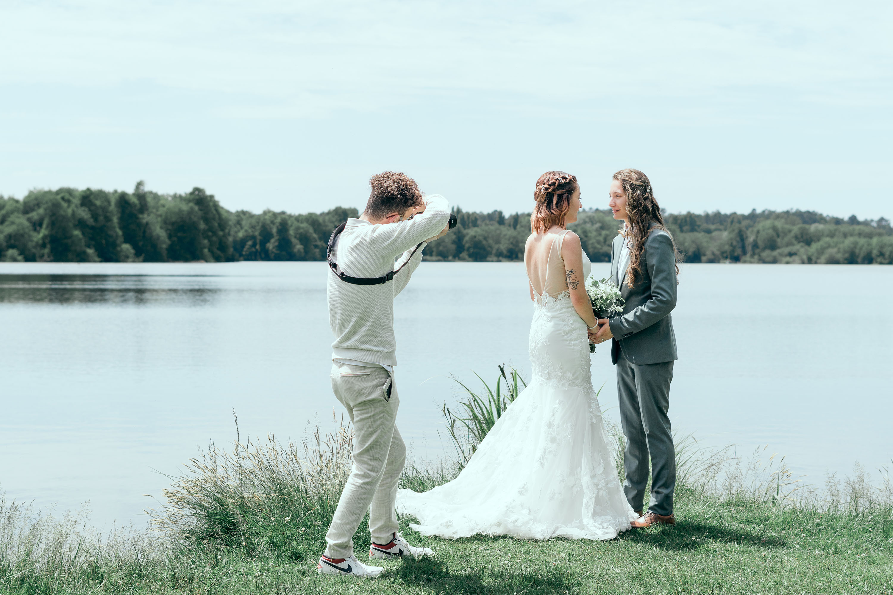 Photographe prenant en photo un couple de mariés au bord d'un lac avec des arbres en arrière-plan